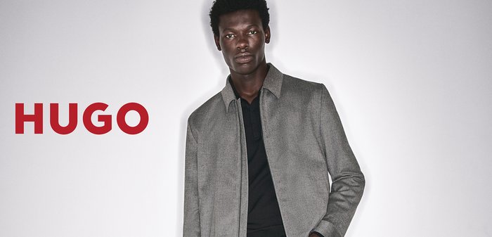 Man wearing grey jacket and jeans holding black bag, standing beside black chair, with red HUGO text on white background.