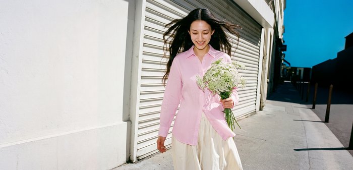 Jeune femme en chemise rose et jupe blanche marche dehors en tenant un bouquet de fleurs blanches, souriante, les cheveux flottant dans la brise.