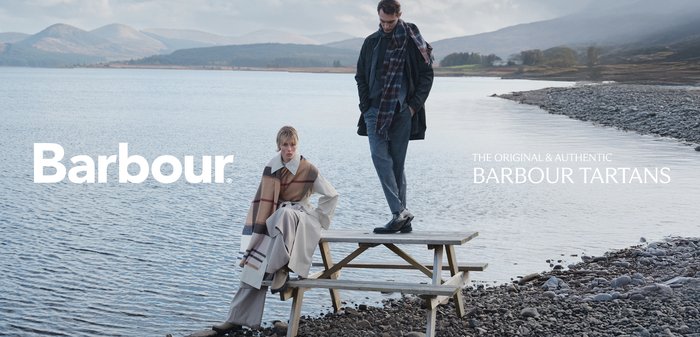 Zwei Models an einem See, die Barbour-Bekleidung tragen. Eines trägt einen karierten Poncho und Hosen, das andere in einem dunklen strukturierten Mantel und Schal.