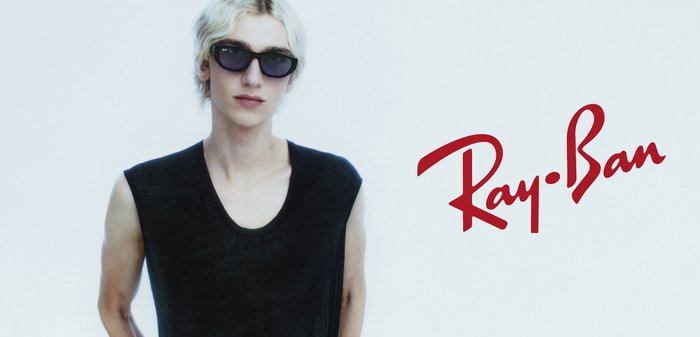 Młoda osoba z krótkimi blond włosami nosi czarne okulary przeciwsłoneczne Ray-Ban i czarny bezrękawnik na jednolitym jasnym tle.