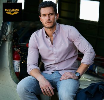 Man draagt een lichtroze overhemd met knopen en spijkerbroek, zit in een vliegtuigloods vlak bij een brandblusser en kijkt bedachtzaam opzij.