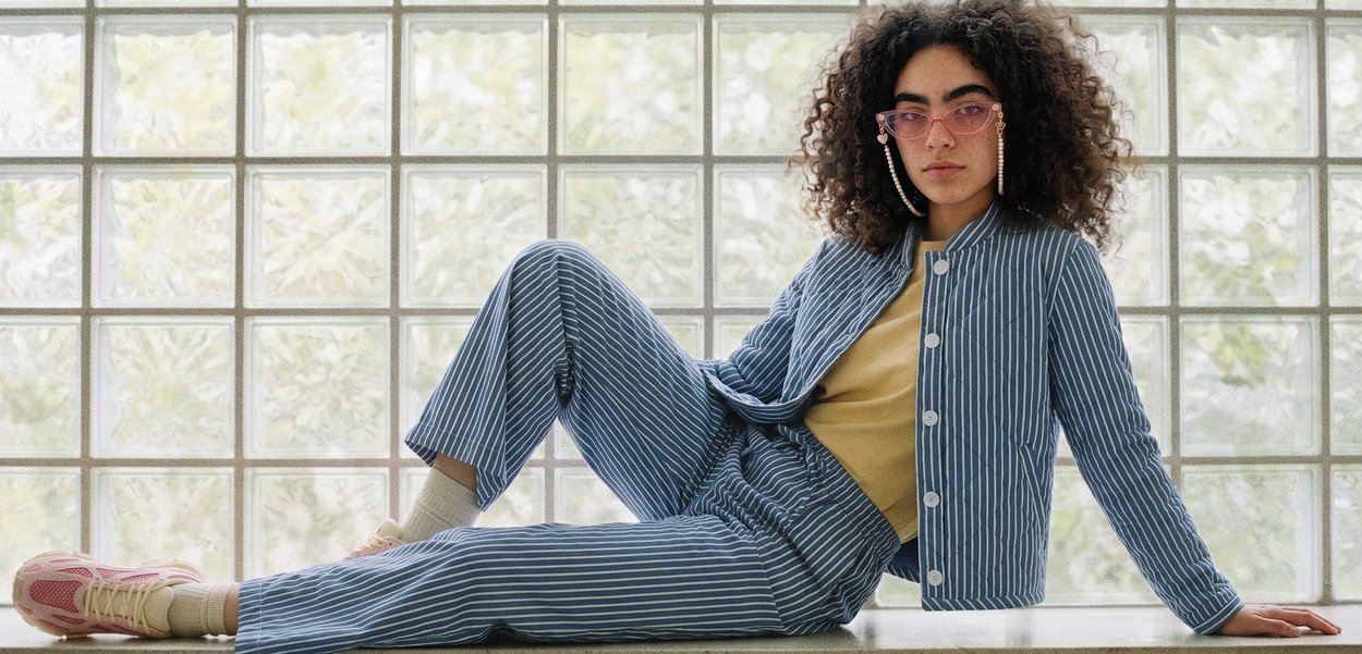 Persona joven con cabello rizado y gafas rosas reclinada en un saliente, vistiendo chaqueta y pantalones azul a rayas con camisa beige y zapatillas.