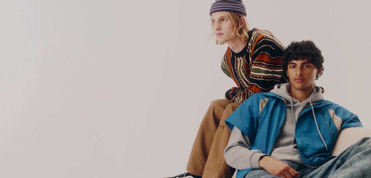 Două persoane stau coteț la coteț. Una poartă un pulover cu un model multicolor și pantaloni maronii; cealaltă este îmbrăcată într-o jachetă albastră peste un hanorac gri.