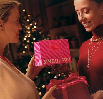 Borsa regalo rosa con la scritta "SINGULARU" in bianco e motivi geometrici, tenuta da una persona e consegnata a un'altra insieme a una scatola regalo rosa.