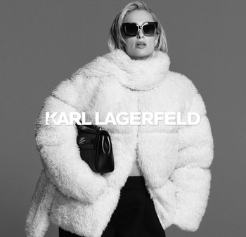 White fuzzy puffer jacket with a high collar, worn over a black top. Model holds a small black bag featuring a subtle logo.