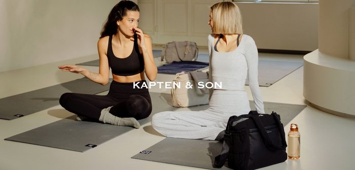 Dva lidé sedí na šedých jógových podložkách. Jeden má na sobě černý krátký top a legíny; druhý světle šedý dlouhý rukáv a odpovídající kalhoty.