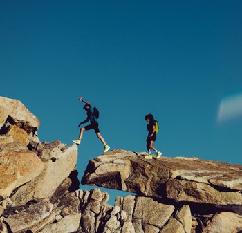 Due persone che navigano su rocce impervie, indossando abbigliamento sportivo nero e calzature gialle vivaci, con piccoli zaini, contro un cielo blu limpido.