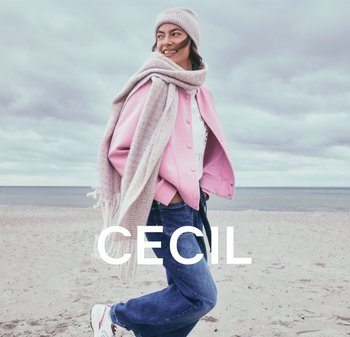 Rosa oversized Jacke mit Knöpfen, weißes Top, blaue Denim-Hosen und ein beiger Strickschal sowie eine Mütze, auf einem sandigen Strand.