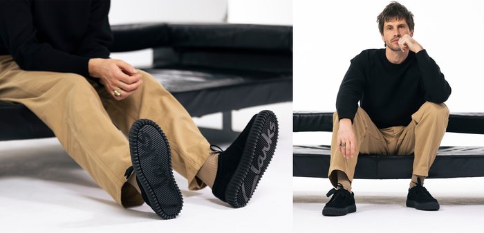 Homme en pull noir et pantalon beige assis sur un banc noir, montrant les semelles de chaussures noires texturées avec la marque « Clarks ».
