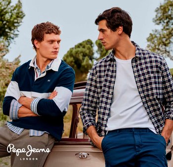 Dos hombres están al lado de un coche vintage. Uno lleva una camiseta de rugby a rayas en azul marino; el otro tiene una camisa de cuadros sobre una camiseta blanca.