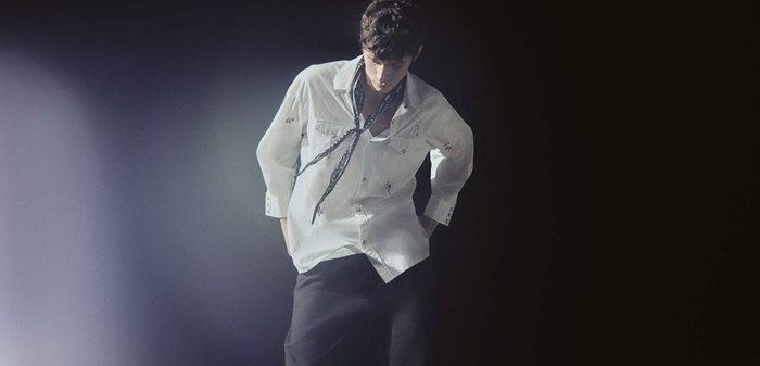 Junger Mann trägt ein weißes, besticktes Hemd mit Knöpfen, schwarze Hose und ein gemustertes Halstuch, steht vor einem dunklen Farbverlauf-Hintergrund.