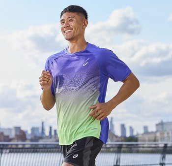 Man joggend buiten op een pad langs het water, gekleed in een paars-groen sportshirt en zwarte korte broek, met een stadsgezicht op de achtergrond.