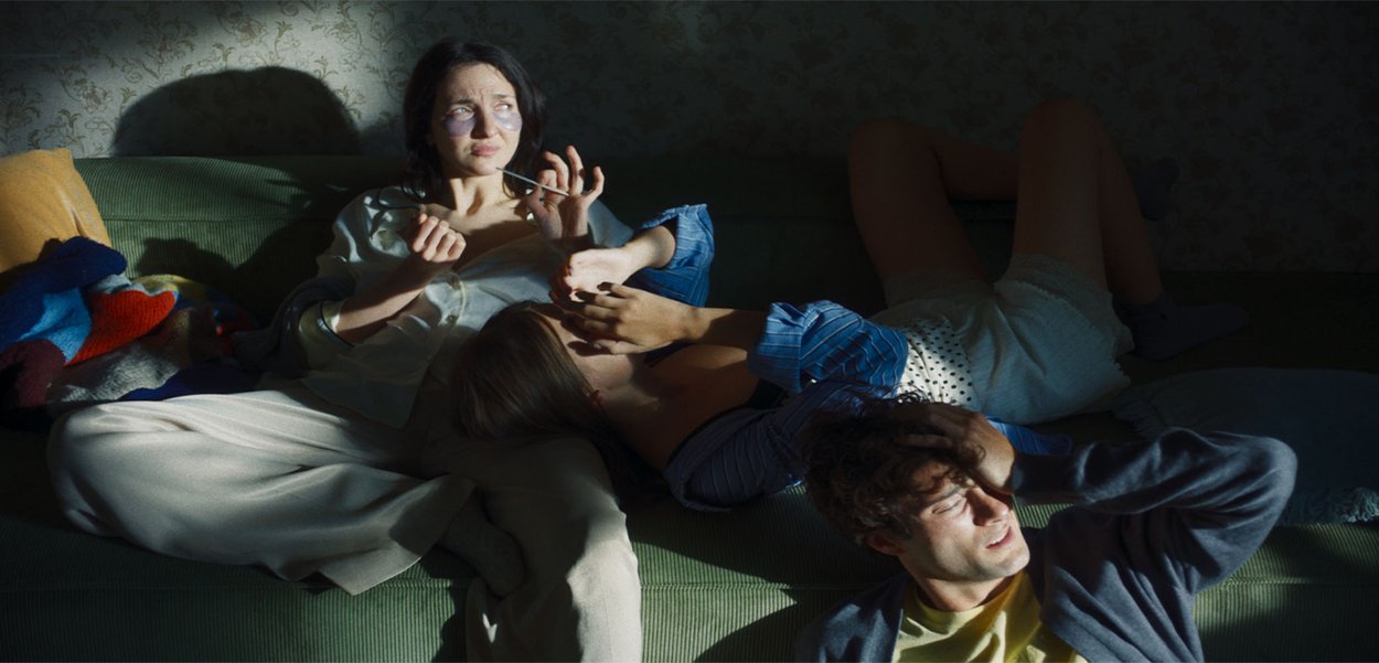 Un divano verde con texture e una coperta colorata. Tre persone sono sdraiate in abiti casual. La scena è illuminata dolcemente, con un'enfasi sulle ombre.