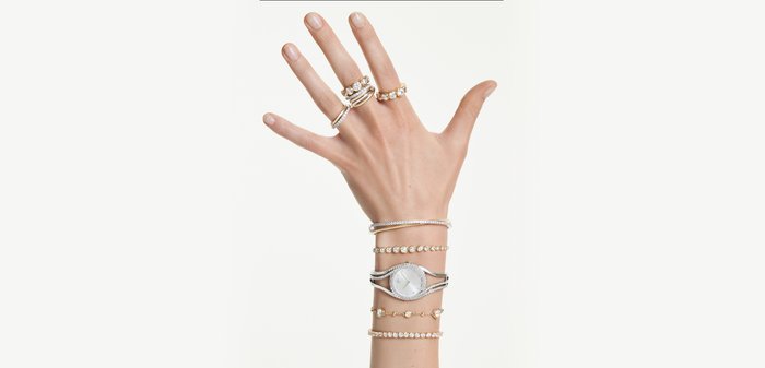 Hand with five gold and diamond rings on fingers and five diamond-studded bracelets, including a silver watch, on wrist against white background.