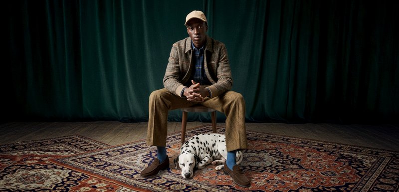 Homme assis sur une chaise en bois, portant un blazer marron à motifs par-dessus une chemise bleue, un pantalon beige et une casquette beige. Un Dalmatien endormi se trouve à proximité.