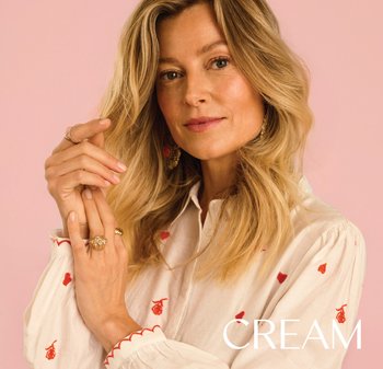 Blonde Frau mit welligem Haar trägt eine weiße Bluse mit roter Stickerei und goldenen Schmuck und posiert vor einem rosa Hintergrund.