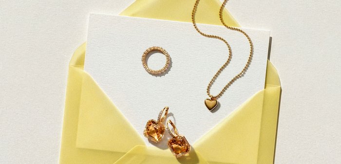 Κίτρινος φάκελος που κρατάει μια λευκή κάρτα με χρυσό κολιέ με καρδιά, χρυσό δαχτυλίδι με πέτρες και χρυσά σκουλαρίκια με κεχριμπαρένιους πολύτιμους λίθους.
