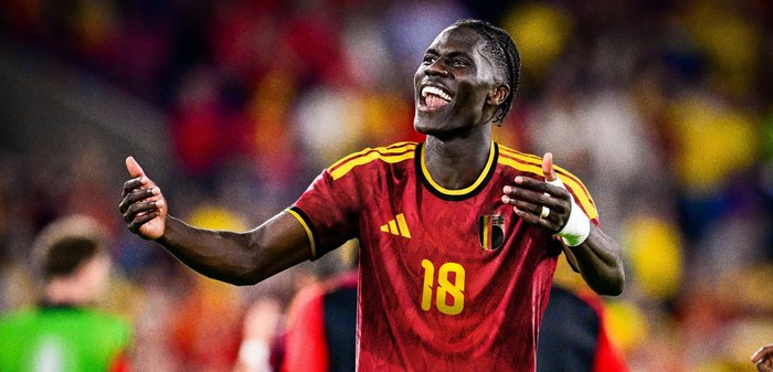Joueur de football belge portant le maillot rouge numéro 18 célèbre sur le terrain avec les bras étendus et un large sourire.