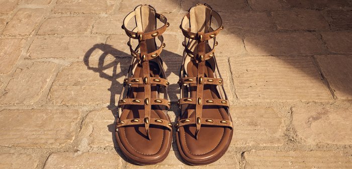 Sandalias gladiador de cuero marrón con tiras al tobillo y tachuelas metálicas, colocadas sobre un pavimento de piedra bajo la luz del sol.