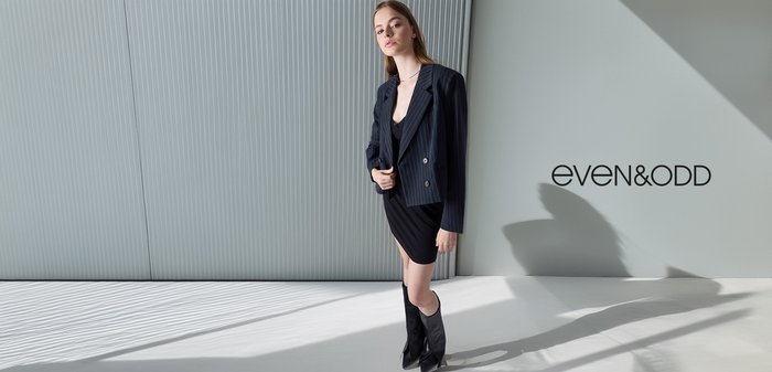 Zwarte jurk met een getailleerde silhouet, gelaagd onder een donker gestreepte blazer. Kniehoge zwarte laarzen, gladde texturen en minimalistische ontwerpelementen.