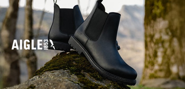 Bottines Chelsea en cuir noir avec des panneaux latéraux élastiques, bout rond et semelle en caoutchouc texturé, posées sur un rocher recouvert de mousse dans la nature.