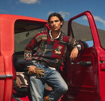 Maglione lavorato a maglia con motivi rossi, blu e verdi, caratterizzato da un orsetto; indossato con jeans di denim e guanti marroni; appoggiato su un veicolo rosso.