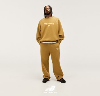 Brązowa bawełniana bluza z białym napisem, w zestawie z dopasowanymi luźnymi spodniami dresowymi. Czarne buty dopełniają strój. Prosty design, luźne dopasowanie.