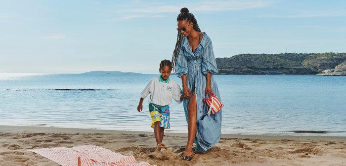 Žena v modrých šatách drží za ruky dieťa, ktoré kráča bosé po piesočnatej pláži pri pokojnom mori pod jasnou oblohou.