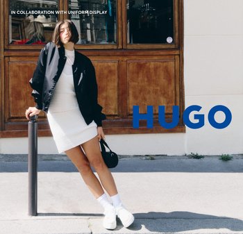 Veste bomber noire sur une robe en tricot blanche ; baskets et chaussettes blanches ; debout à côté d'un poteau devant une fenêtre en bois. Texte bleu "HUGO".
