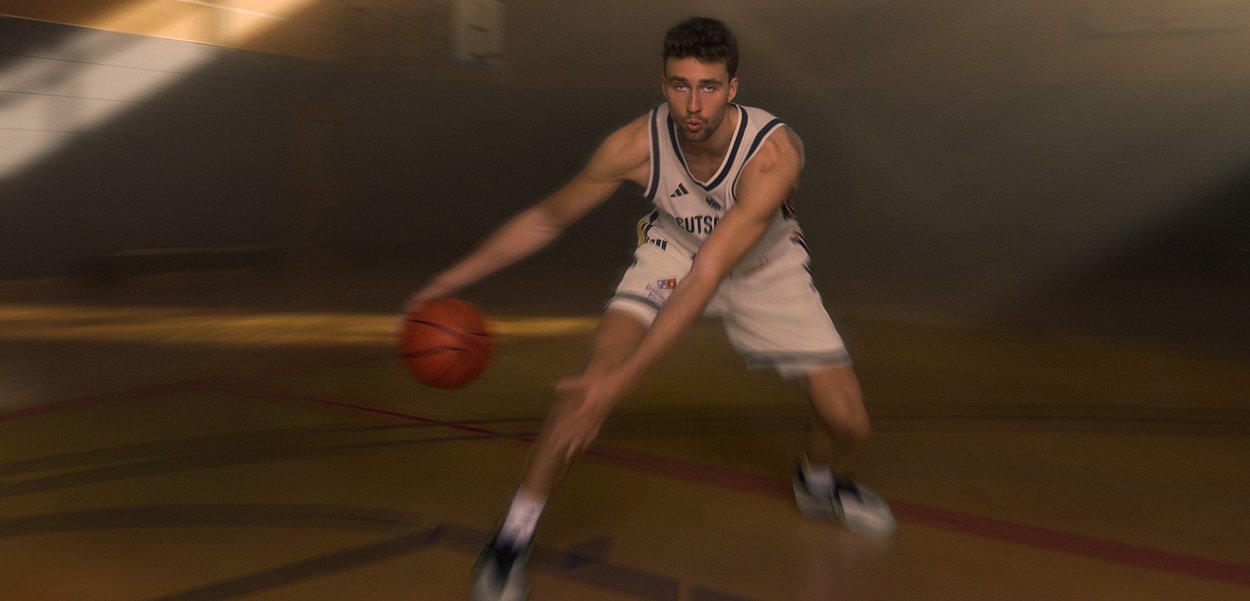 Jugador de baloncesto masculino driblando un balón cerca del suelo en una cancha cubierta con expresión concentrada.