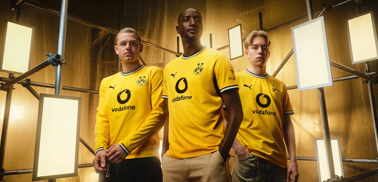 Drei Männer in gelben Fußballtrikots von Borussia Dortmund stehen selbstbewusst zwischen hellen, rechteckigen Studiolichtern und einem goldenen Hintergrund.