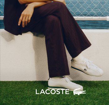 Persona sentada en un borde blanco junto a una valla con red, vistiendo pantalones oscuros y zapatillas blancas Lacoste sobre césped verde. Se ve el logo de Lacoste.