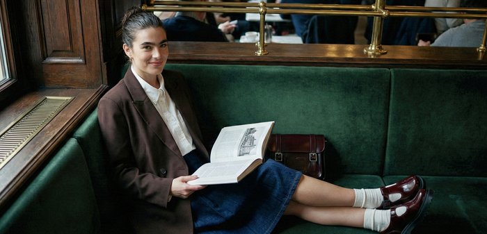 Jeune femme assise sur un banc en velours vert près d'une fenêtre, lisant un livre, portant un blazer marron, une jupe, des chaussettes blanches et des chaussures foncées.