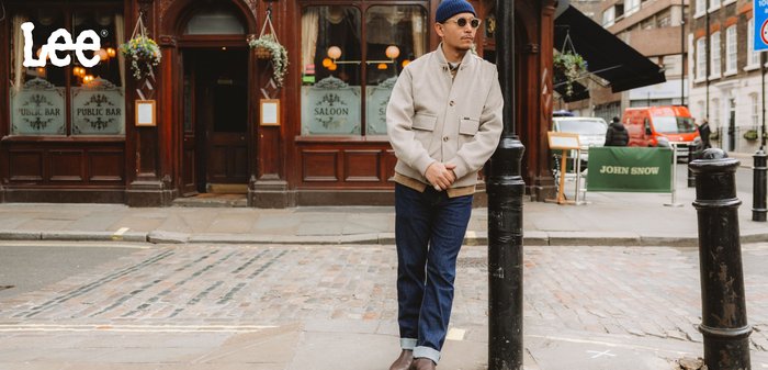 Ljusgrå jacka i ullblandning med framfickor, beige detaljer, blå jeans och bruna stövlar, poserande mot en bakgrund av en pub.