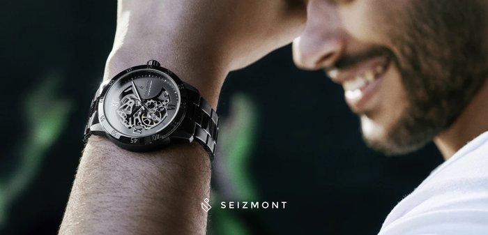 Black skeleton watch with metallic accents, round face, clear display of gears, and linked bracelet design, worn on a wrist.
