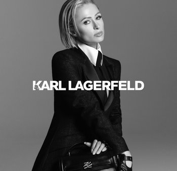 Traje negro a medida con un cuello de camisa blanca; cabello corto y claro; sosteniendo un bolso negro con las iniciales "KL" en un diseño minimalista.