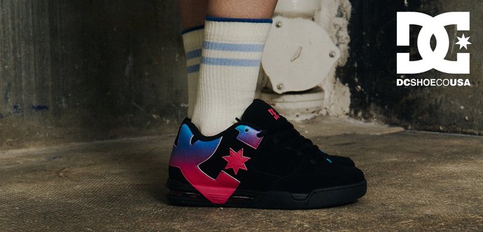 Schwarze Sneaker mit pinken und blauen Akzenten, kombiniert mit weißen Socken mit blauen Streifen, getragen von einem sitzenden Model in einem Jeansrock.