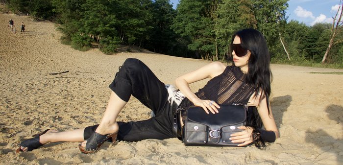Black leather crossbody bag with rounded edges, two front pockets, and silver hardware. Model wears a black mesh top and loose black pants.