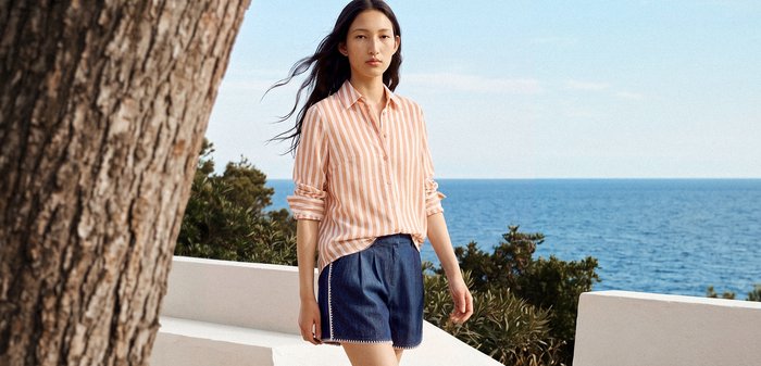Jeune femme aux longs cheveux bruns portant une chemise rose à rayures et un short bleu, debout près d’un mur blanc au bord de la mer par une journée ensoleillée.