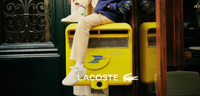 Child wearing a navy hoodie with green crocodile logos, beige trousers, and white trainers sitting on a yellow postbox next to a dark green door.