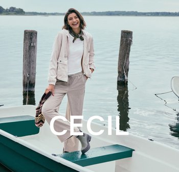 Lichtbeige zip-up jacket met trekkoorden, witte top, grijze wijde broek en grijze regenlaarzen; staand op een boot aan het water.