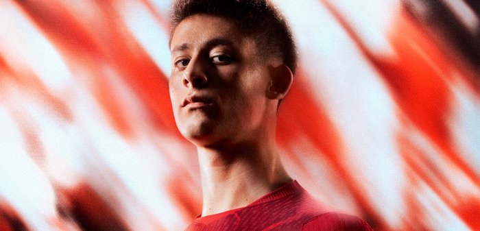 Jeune homme aux cheveux courts, portant une chemise rouge, regardant la caméra avec confiance sur un fond flou rouge et blanc.