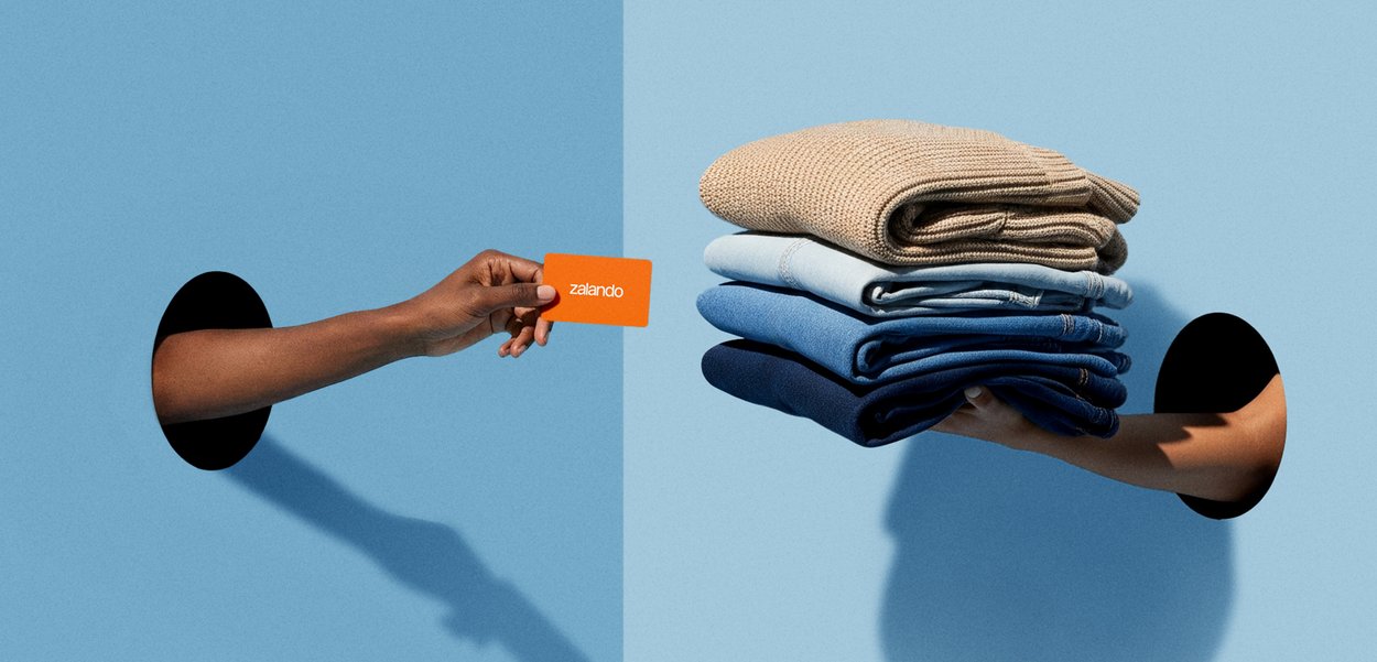 Zalando-presentkort hålls i en brun hand, mittemot en hög med olika blå stickade och vävda tröjor, presenterade av en annan hand genom ett hål.