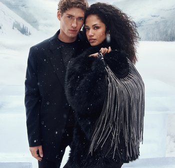 Ytterkläder av svart päls med silverfransar, bärs av modeller i en snöig miljö. En modell håller en pärlbroderad handväska, båda klädda i mörka kläder.