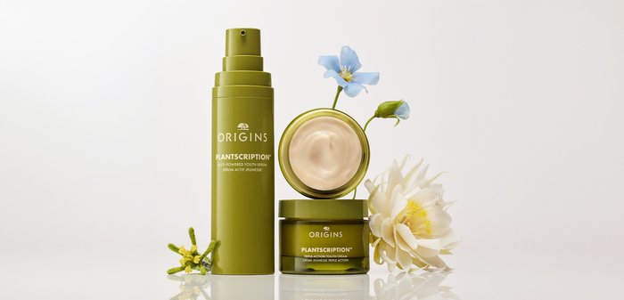 Flacon de sérum Plantscription Green Origins et pot de crème ouvert avec crème beige clair, entourés de petites fleurs bleues et blanches sur surface blanche.