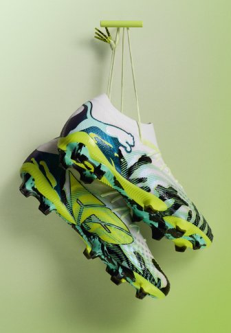 Crampons de football blancs et verts néon avec des accents noirs suspendus par les lacets sur un fond vert clair.