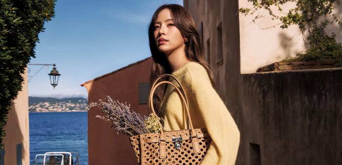 Femme en pull jaune tenant un sac tissé avec des fleurs, debout dans une rue ensoleillée au bord de la mer sous un ciel bleu clair.