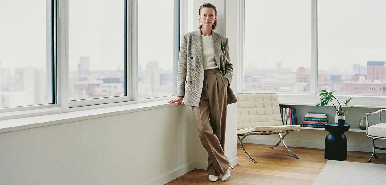 Grå houndstooth-blazer, hvit topp og beige plisserte bukser. Hvite sko. Moderne interiør med tregulv, store vinduer og dekorative planter.