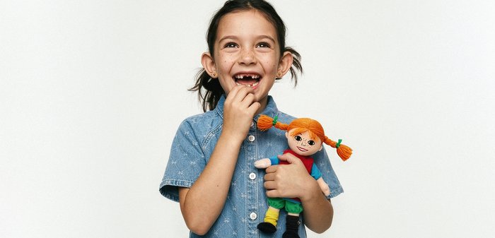 Χαμογελαστή νεαρή κοπέλα με κενά στα μπροστινά δόντια κρατώντας μια κουκλίτσα με κόκκινα μαλλιά και πλεγμένα κοτσίδια, σε λευκό φόντο.