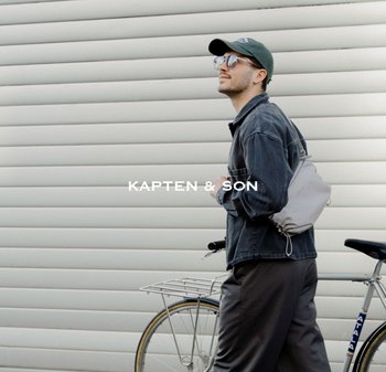 Mladý muž nosící sluneční brýle, čepici a džínovou bundu kráčí vedle kola s světle šedým batůžkem na stažení u bílé zdi.